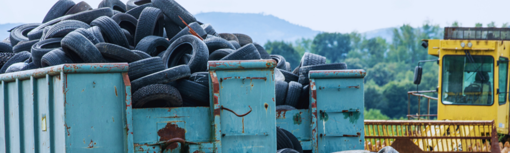 Pile of used tires in a rusted turquoise trailer, with a yellow construction vehicle in the background.