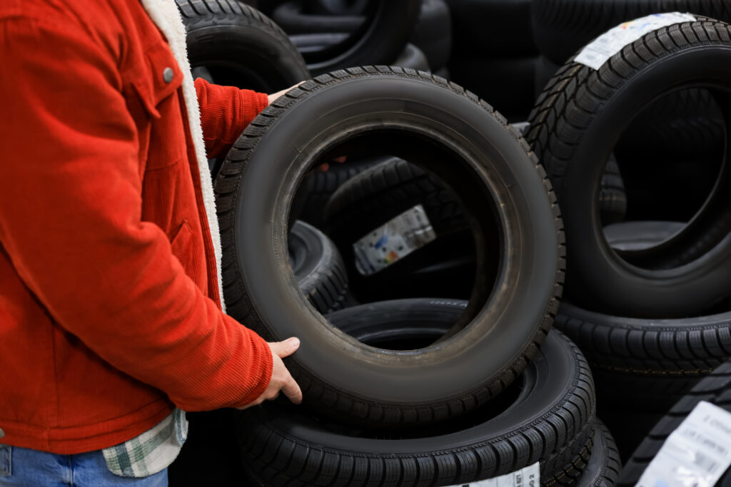 Person in a red jacket holding a tire in a tire shop with stacked tires in the background
