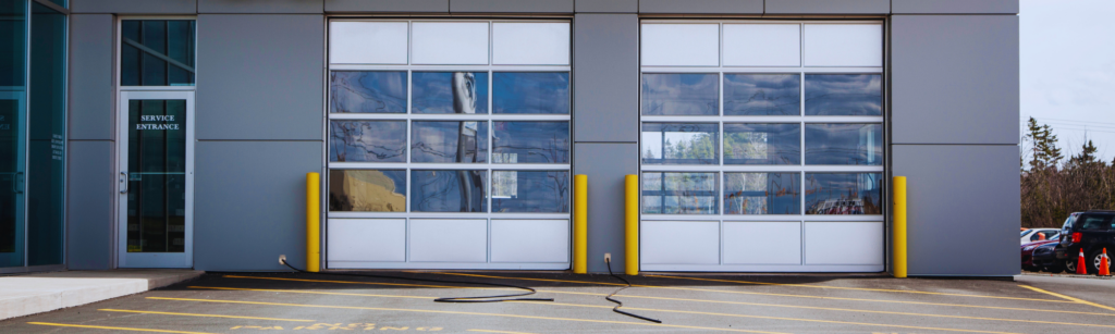 Modern service building with two large glass garage doors flanked by yellow protective bollards; a glass entry door labeled 'Service Entrance' sits to the left, and cars are visible in the parking area to the right. A black hose lies on the asphalt in front of the doors.