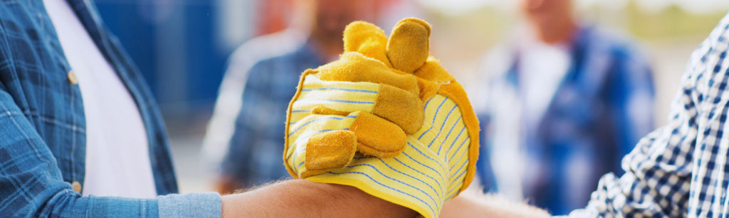Two people wearing yellow work gloves clasping hands in a handshake, with blurred workers in the background, suggesting teamwork on site.
